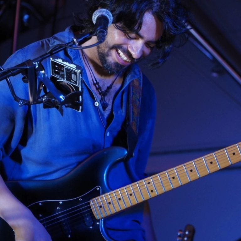 A man with long dark hair and a beard plays an electric guitar on stage in Roatan, Honduras, smiling as he performs under blue stage lighting.