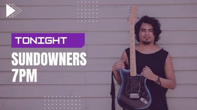A person with curly hair wearing a black sleeveless shirt holds an electric guitar in front of a light-colored wooden wall on Roatan, Honduras.