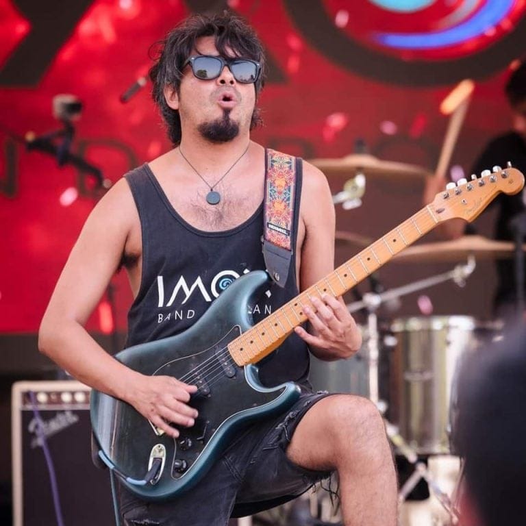 A musician wearing sunglasses and a black tank top with "IMAGINE" printed on it plays an electric guitar during a live performance on Roatan, Honduras, with a red and colorful backdrop and a drummer visible in the background.