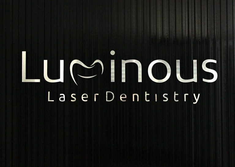 A black vertical panel features a sign with the word "Luminous" in large white letters, incorporating a smile and a curved line resembling a tooth as part of the logo, along with the words "Laser Dentistry" beneath it, indicating a dental clinic in Roatan, Honduras.