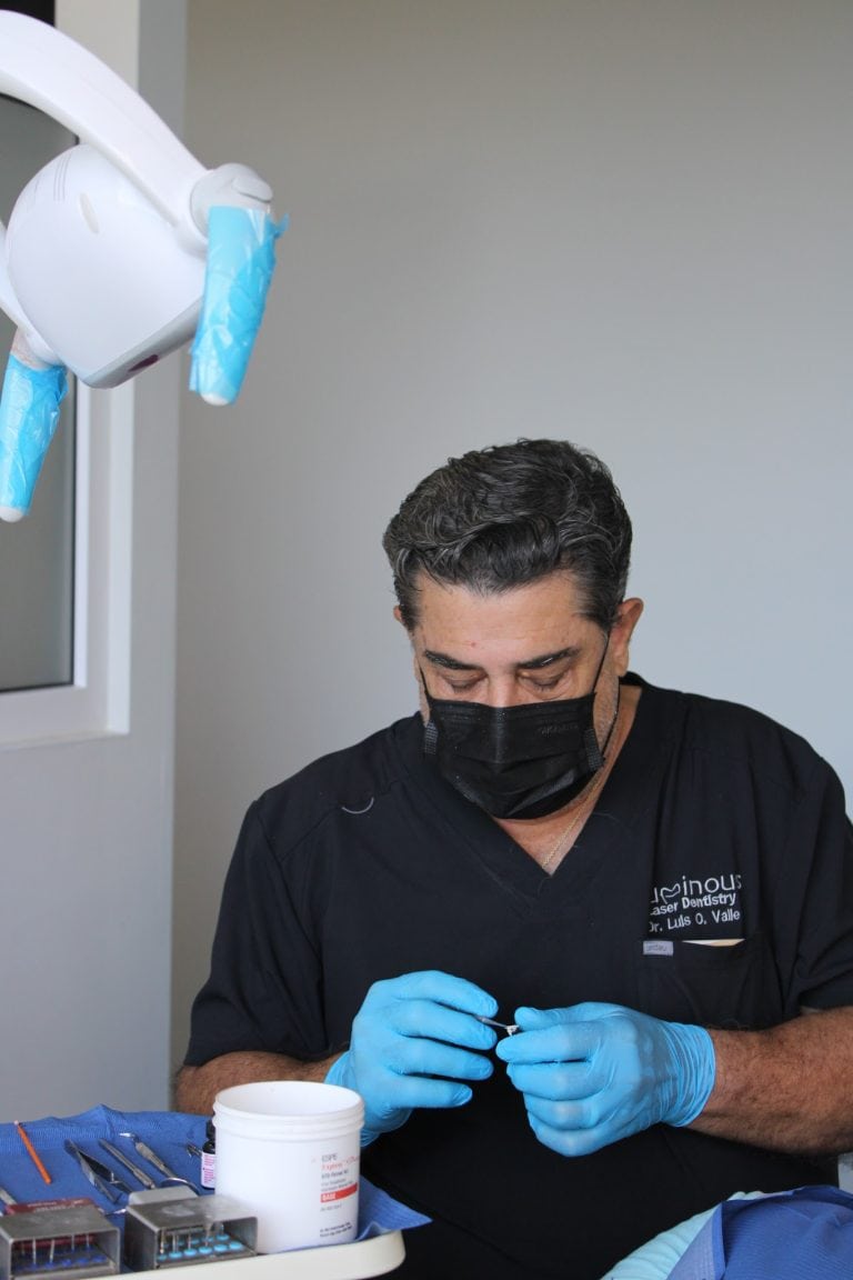 A male dentist in black scrubs and a face mask, wearing blue gloves, prepares dental instruments at a clinic in Roatan, Honduras, with dental tools and supplies arranged on a table.