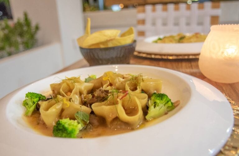 A plate of Asian-style noodles with vegetables and herbs is served on a table with bowls of chips and a lit candle, set against the backdrop of a patio in Roatan, Honduras.