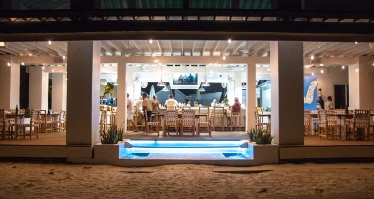 An illuminated beachfront restaurant in Roatan, Honduras features a bar area with seating, string lights, and a large television, with people gathering inside and a sandy beach in the foreground.