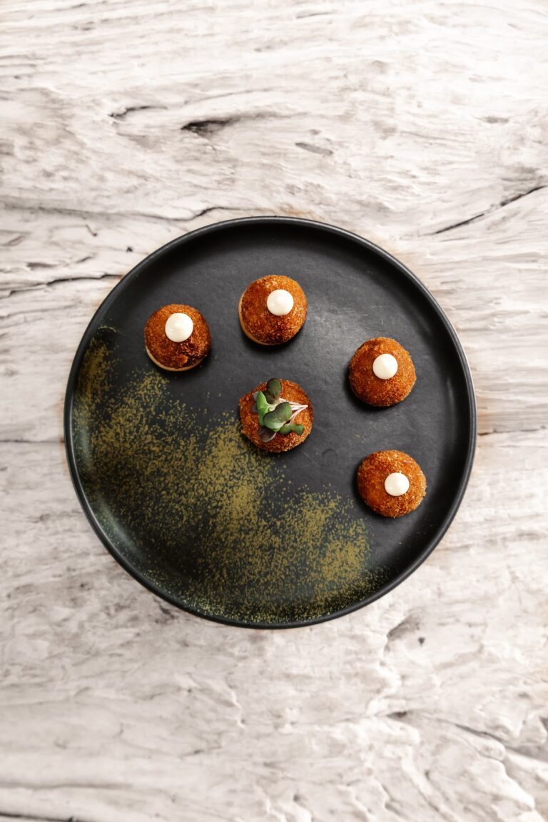 Four crispy appetizer balls topped with white sauce and microgreens are arranged on a black plate with a sprinkle of yellow powder, set on a white marble table in Roatan, Honduras.