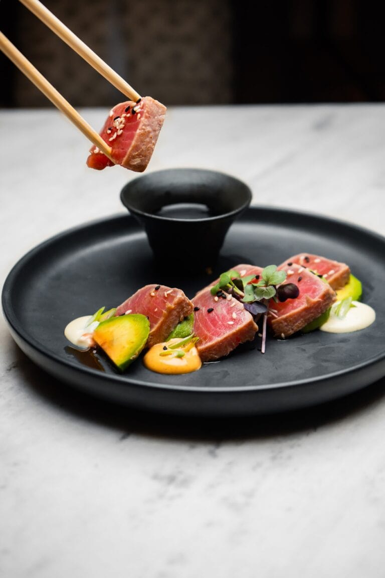 A slice of seared tuna topped with sesame seeds and microgreens is being dipped into a black soy sauce dish on a black plate, set against a white marble surface in Roatan, Honduras.