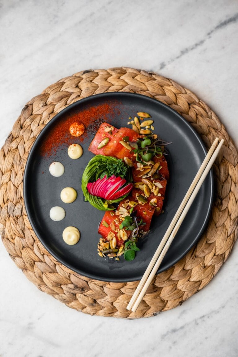 A beautifully plated dish featuring sliced watermelon topped with microgreens, nuts, and edible flowers, served on a black plate with varied sauces, placed on a woven placemat on a white marble surface in Roatan, Honduras.