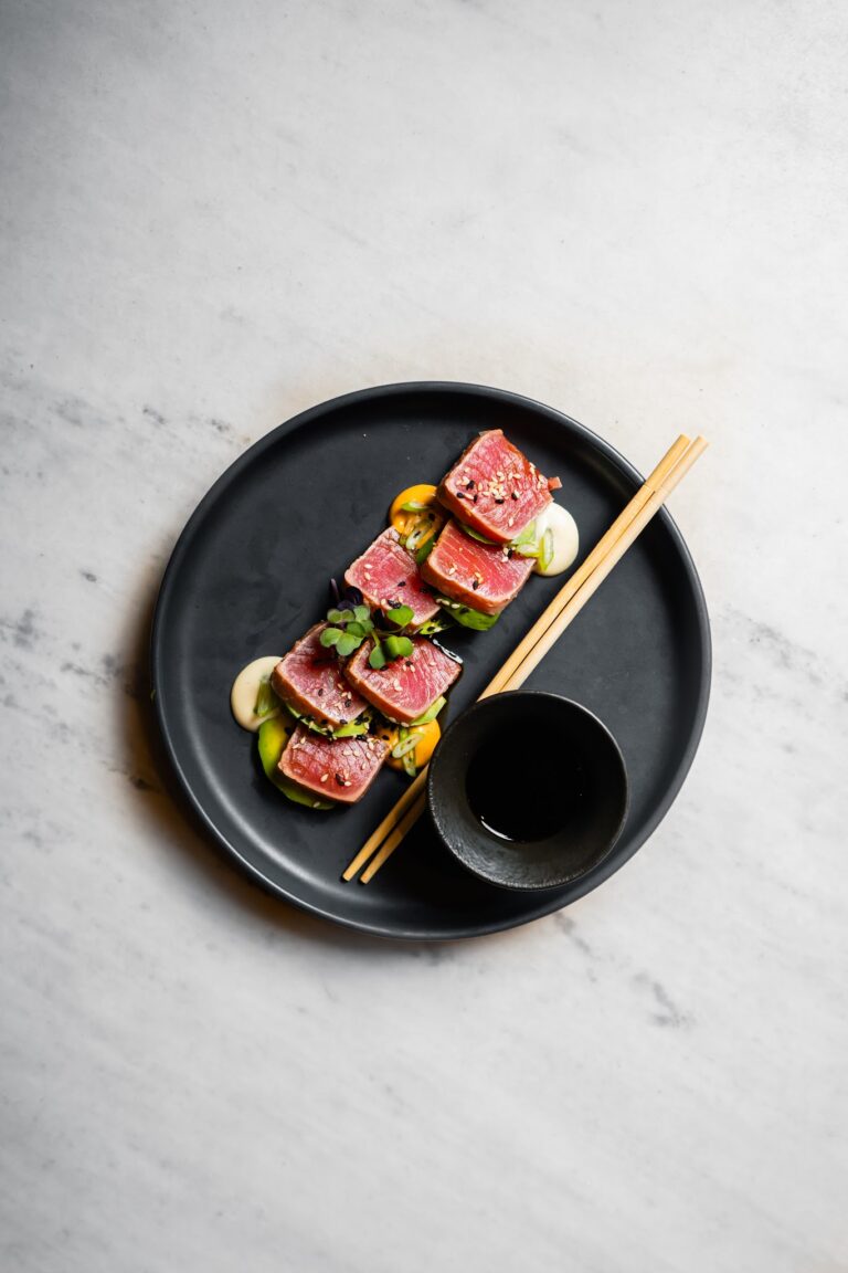 Sliced ahi tuna sashimi garnished with microgreens and sesame seeds served on a black plate with a small dipping bowl of soy sauce and chopsticks on a marble surface, representing a dish available in Roatan, Honduras.