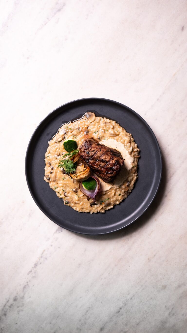 A plated dish featuring grilled meat, creamy risotto with mushrooms, and garnished with herbs and vegetables, served on a black plate with a white marble surface background in Roatan, Honduras.