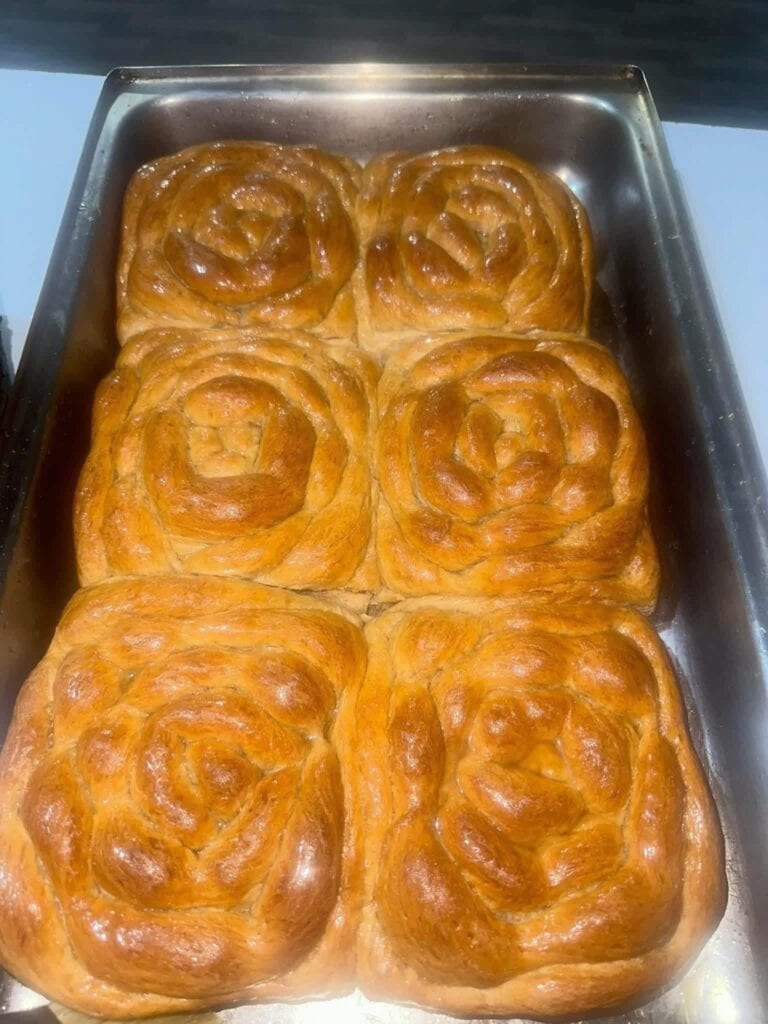 Four freshly baked cinnamon rolls with glossy, golden-brown tops are arranged in a metal baking tray, set against a background indicating a location in Roatan, Honduras.