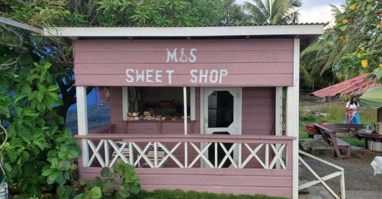 A small pink wooden shop labeled "MSS Sweet Shop" is situated on Roatan in Honduras, with baked goods displayed inside and surrounded by lush tropical greenery and outdoor seating.