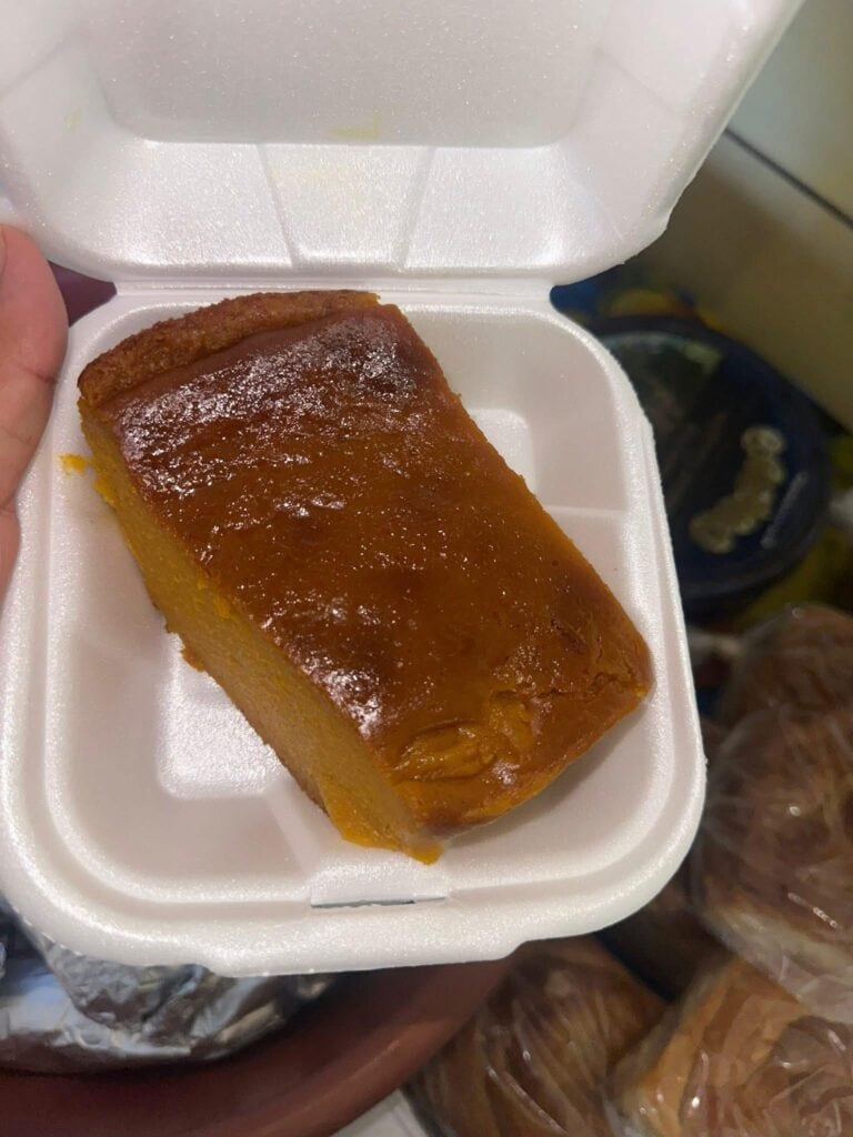 A slice of caramelized flan sits in a white foam container, with a slightly glossy surface and light dusting of powdered sugar, set against a background of various plastic-wrapped food items on a market or street vendor display in Roatan, Honduras.