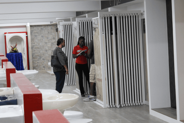 Three people are inspecting shower stalls in a modern bathroom showroom on Roatan, Honduras, with white showers, a stone accent wall, and a display in the background.