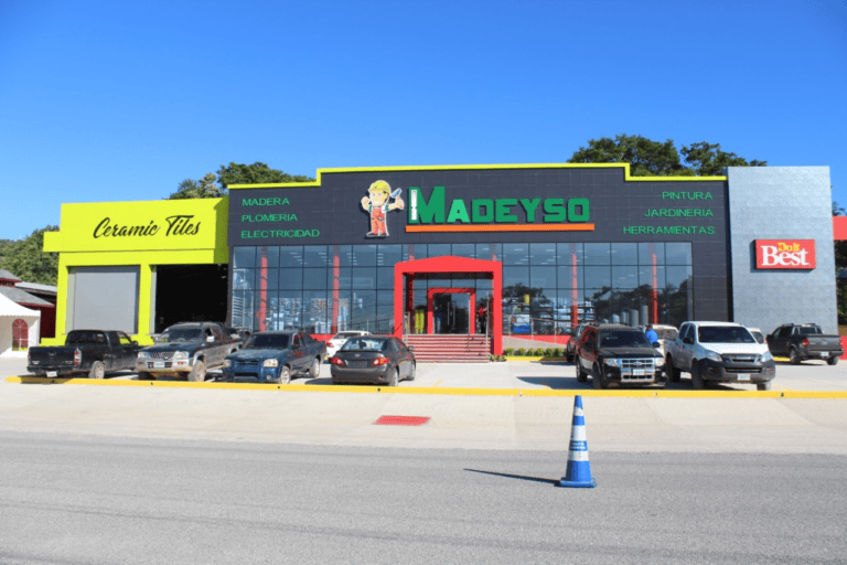 A commercial building in Roatan, Honduras, with signage for Madeyso hardware store, ceramic tiles, and Doit Best, featuring large glass windows, a red entrance, and a parking lot with multiple parked vehicles under a clear blue sky.