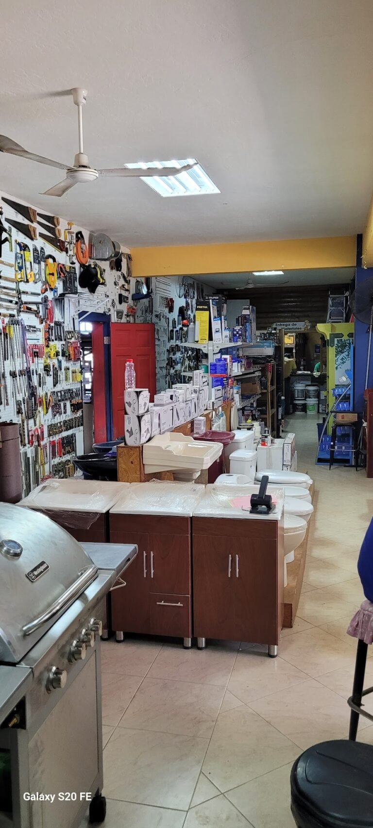 An interior view of a hardware store in Roatan, Honduras, showcasing shelves stocked with tools, building materials, and household supplies, with a section of toilets and a barbecue grill visible in the foreground.