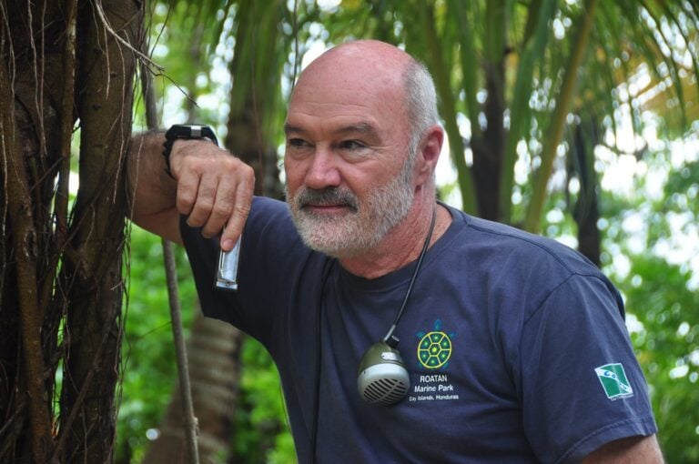 A man with a gray beard wearing a navy blue T-shirt with a Roatan Marine Park logo and a walkie-talkie around his neck stands outdoors among tropical trees in Roatan, Honduras.