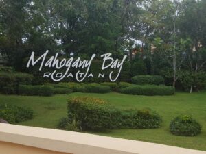 A large sign reading "Mahogany Bay Roatan" is situated among lush green trees and neatly trimmed bushes in Roatan, Honduras.