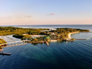 An aerial view of a coastal area in Roatan, Honduras, showing lush green islands, a sandy beach, marinas, and calm blue waters under a clear sky.