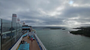 A view from the deck of a cruise ship sailing near Roatan, Honduras, showing the ship's seating area with blue lounge chairs and passengers, with an expansive ocean and small islands under a cloudy sky in the background.