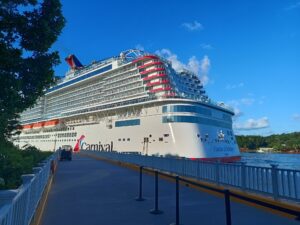 A large white Carnival cruise ship named Carnival Jubilee is docked in Roatan, Honduras, with a walkway and white fencing leading up to it against a bright blue sky.