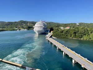 A large white cruise ship docked at a pier surrounded by lush green hills on Roatan, Honduras, with calm blue waters and a clear sky overhead.