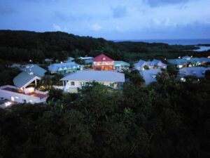 Colorful houses with metal roofs are situated among lush green trees on a hillside near the ocean in Roatan, Honduras, with a forested background and a partly cloudy evening sky.