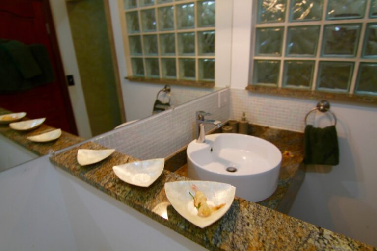 A bathroom in a business on Roatan, Honduras, features a white vessel sink, a granite countertop with decorative shell-shaped bowls, and large glass block windows allowing natural light to fill the space.