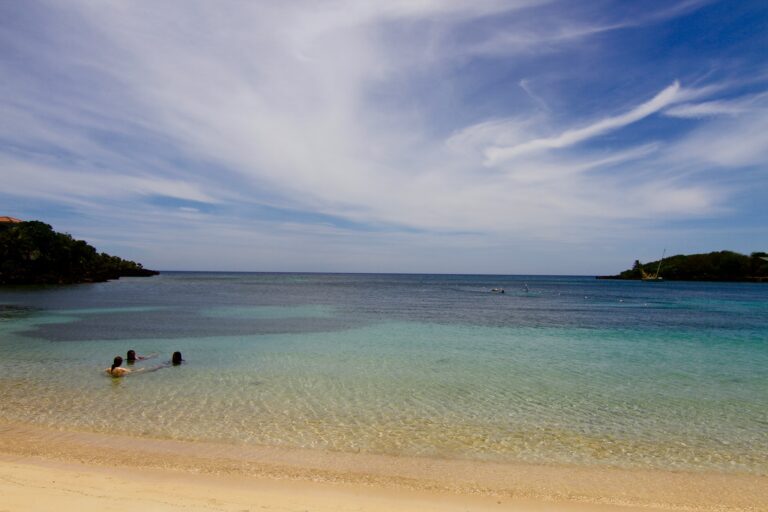 A clear view of Roatan's white sandy beach with calm turquoise waters, a few people swimming, and lush green islands under a partly cloudy sky in Honduras.
