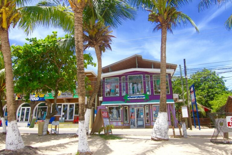 Colorful storefronts and palm trees line a sandy street in Roatan, Honduras, with shops displaying clothing and signs amidst lush tropical greenery.