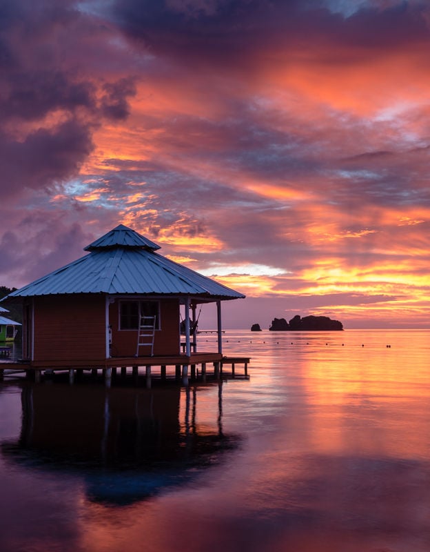 A wooden hut with a metal roof sits on stilts above calm Caribbean waters during a vibrant sunset in Roatan, Honduras, with colorful clouds and an island silhouette in the background.