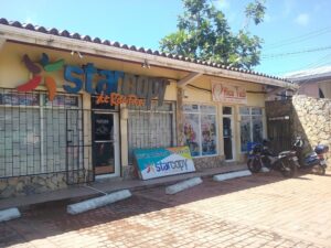 A storefront in Roatan, Honduras, displays signage for "StarCopy de Roatan," with a yellow building façade, colorful logo, and promotional posters in the windows, with motorcycles parked in front and a tree and power lines visible against a clear blue sky.