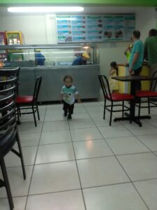 A young boy walking through a casual fast-food restaurant in Roatan, Honduras, with a food counter, menu boards, and several customers standing and waiting nearby.