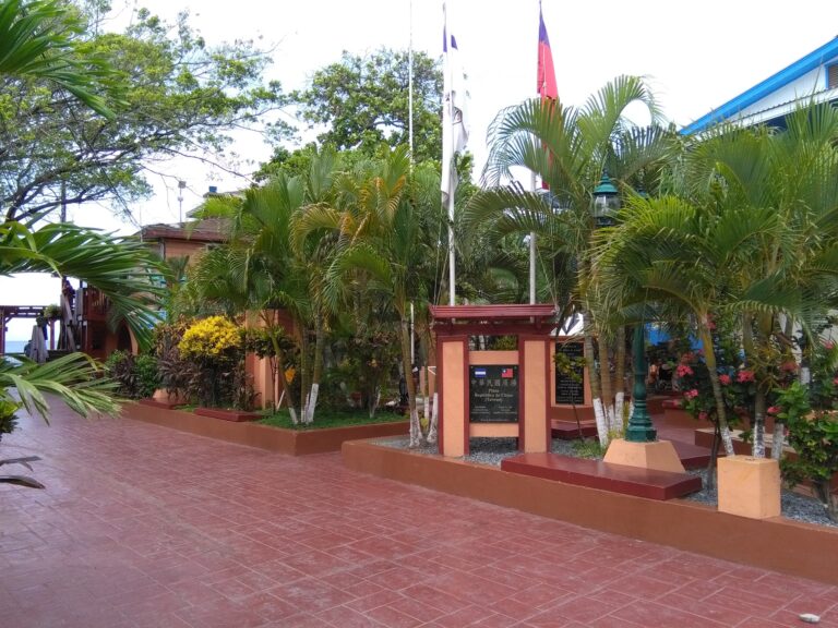 A landscaped area in Roatan, Honduras, features tropical plants, flagpoles with flags, a sign for the Republic of China (Taiwan), and a brick pathway, with a building partially visible in the background.