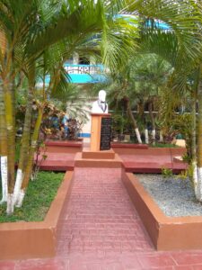 A landscaped courtyard in Roatan, Honduras features a statue of a man in a suit on a pedestal surrounded by lush green palm trees, with a brick pathway leading up to it.