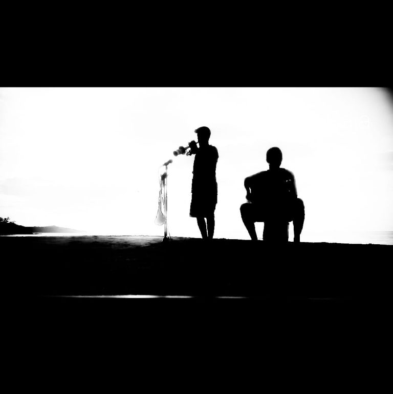 Silhouettes of two people, one standing and playing a guitar and the other sitting, photographed on a beach in Roatan, Honduras, with a bright sky and distant shoreline visible in the background.