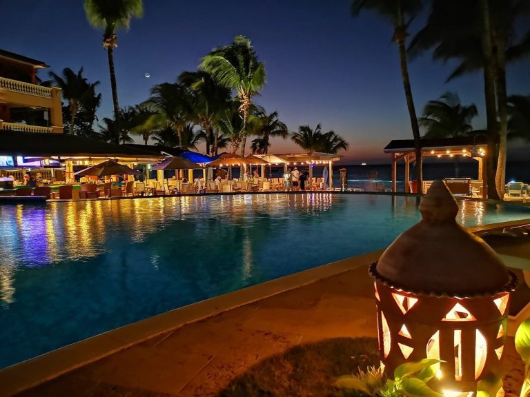 A nighttime scene at a beachside pool in Roatan, Honduras, featuring illuminated seating areas, palm trees, and the ocean in the background.