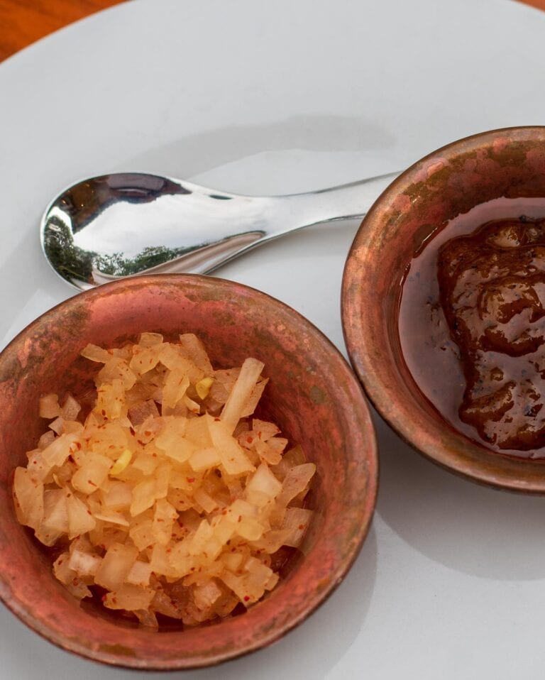 Small copper bowls containing diced onions and a savory sauce are placed on a white plate at a restaurant in Roatan, Honduras.