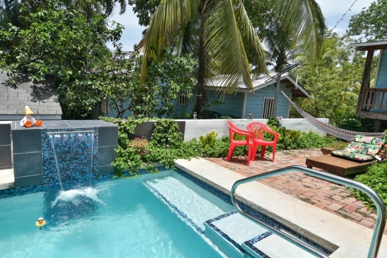 A backyard swimming pool area with a waterfall feature, surrounded by tropical trees, colorful chairs, a hammock, and small houses, on Roatan in Honduras.