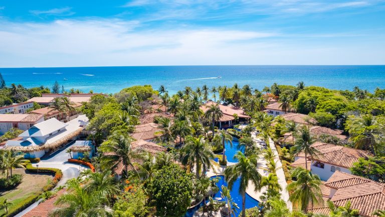 Aerial view of a tropical resort with red-tiled roofs, lush greenery, and a large swimming pool overlooking the turquoise waters of Roatan, Honduras, with boats cruising on the ocean in the background.