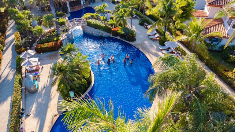 Aerial view of a tropical resort in Roatan, Honduras, featuring a large swimming pool with people enjoying the water, surrounded by lush palm trees, colorful flowering bushes, lounge chairs with umbrellas, and nearby white buildings with terracotta roofs.