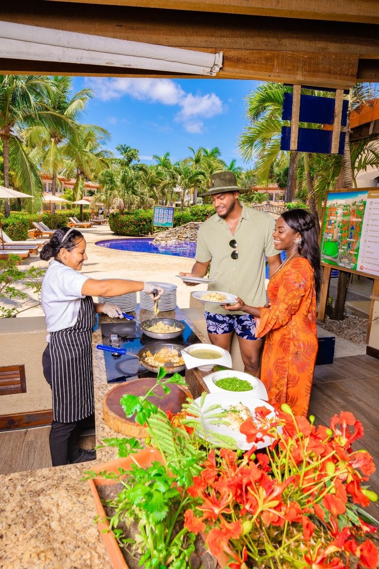 Tourists enjoying a delicious meal at an outdoor restaurant in Roatan, Honduras, with lush tropical palm trees, a swimming pool, and vibrant flowers in the background.