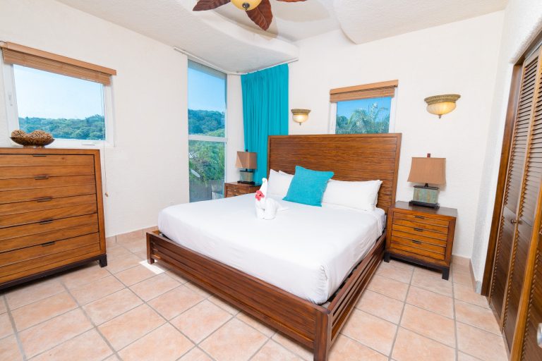 A bedroom in a Roatan, Honduras hotel features a wooden bed with white linens and a blue accent pillow, flanked by matching wooden nightstands with lamps, large windows with blinds and blue curtains, and tropical greenery visible outside.