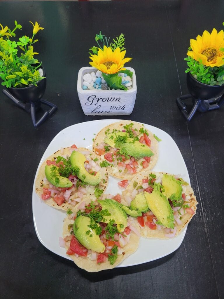Three fish tacos topped with avocado, onion, tomato, and cilantro are served on a white plate, with decorative yellow sunflowers in black pots and a white container with a sunflower and the words "Grown with Love" on a dark table in Roatan, Honduras.