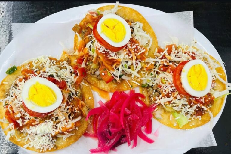 Three crispy tostadas topped with shredded cheese, chopped vegetables, sliced tomatoes, and halved boiled eggs are served on a white plate with pickled pink onions in the center, set on a dark table in Roatan, Honduras.
