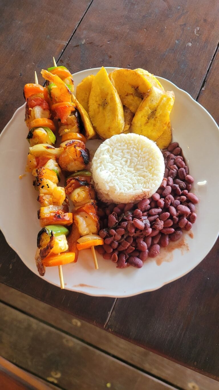 A plate of traditional Honduran food on a wooden table in Roatan, Honduras, featuring skewers of grilled meat and vegetables, fried plantains, red beans, and a mound of white rice.