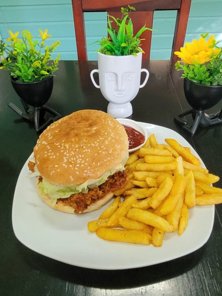 A plate of fish sandwich with French fries, ketchup, and lemon wedges is placed on a black table, with green potted plants and a decorative face-shaped white vase behind it, set against a turquoise and wooden wall in Roatan, Honduras.