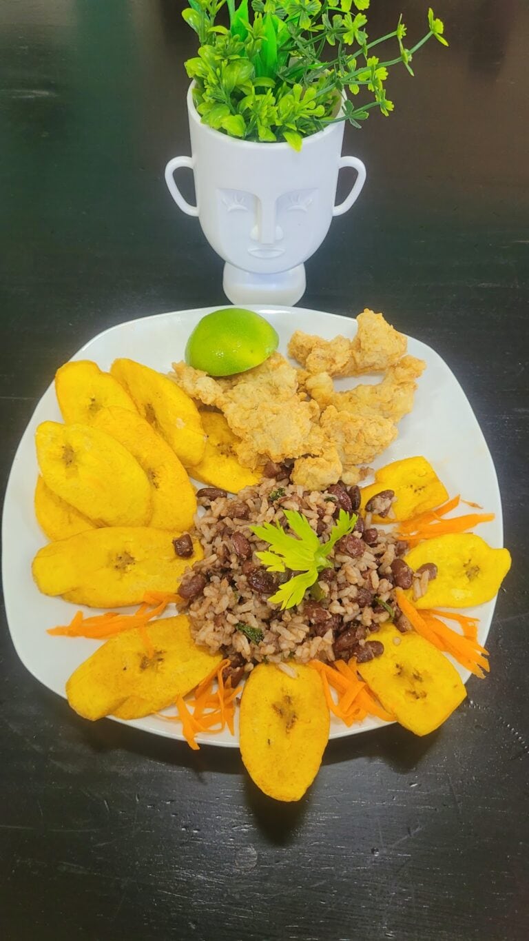 A plate of traditional Honduran food served on Roatan features fried plantains, black beans and rice, shredded carrots, and a lime wedge, with a decorative white ceramic face-shaped planter with green plants in the background.