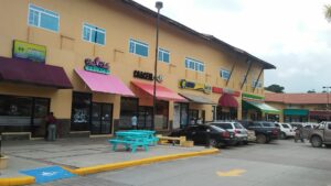 A shopping plaza in Roatan, Honduras, features various storefronts with colorful awnings and people walking in the parking lot, including shops like Costa Dulce Gelato and a cooperative store, with several parked cars and a bright blue picnic table in the foreground.