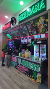 A colorful Mexican food stand in Roatan, Honduras, features neon signs, a menu with tacos, nachos, and tortas, and customers ordering while a staff member prepares food behind the counter.