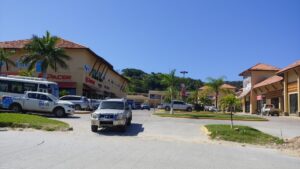 A parking lot in a commercial area on Roatan, Honduras, with several cars, tropical palm trees, and a large shopping center with multiple storefronts and signage against a backdrop of green hills and a clear blue sky.