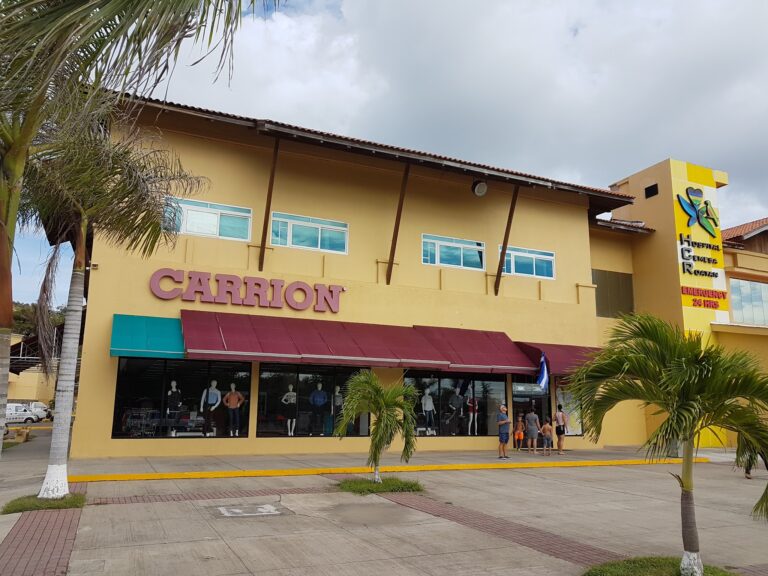 A yellow commercial building on Roatan, Honduras, features a sign for Carrion and a healthcare facility sign for Hospital CEMESA with an emergency 24-hour service, with palm trees and people walking in front.